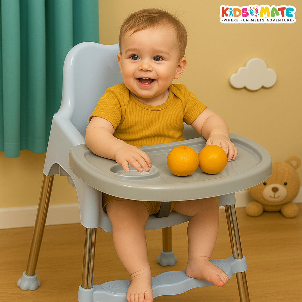 A cheerful toddler sits in a light blue high chair with a gray tray, holding two oranges. The background features a playful, colorful decor, enhancing the joyful atmosphere.