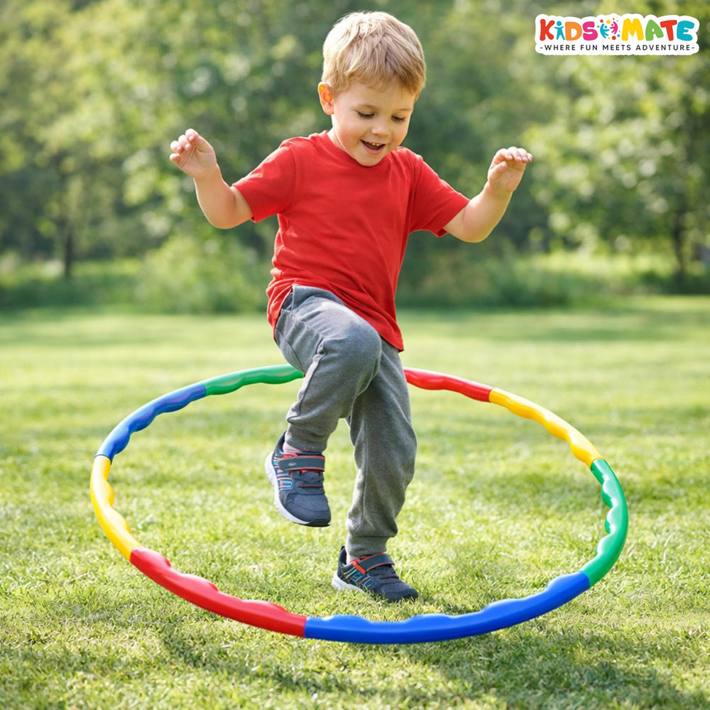 A joyful child in a red shirt and gray pants is playfully jumping through a colorful adjustable hula hoop on a grassy field. The hoop features vibrant blue, red, yellow, and green sections.