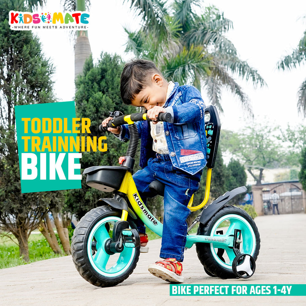 A young child riding a colorful balance cycle in a park setting. The bike features a bright yellow frame with teal accents. The child is smiling, wearing a denim jacket and blue jeans, surrounded by greenery.